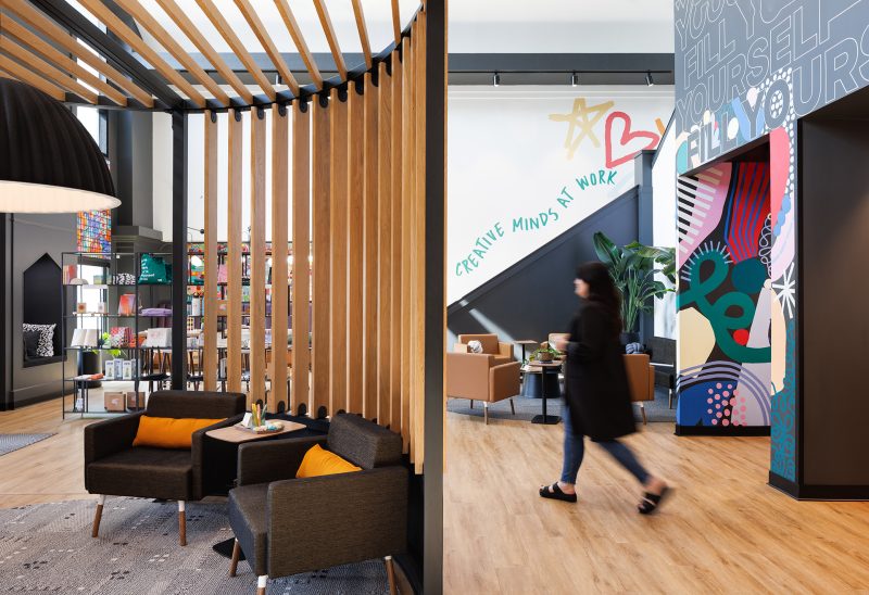 On the left of the Penzone Base One lounge there is a wood, slatted structure with hanging acoustic pendant lamp, dark grey armchairs with yellow pills and a round black coffee table with books and pants on top. On the right is a seating area with brown modern arm chairs and a colorful graphic mural on the wall. A woman in black walks through the space.