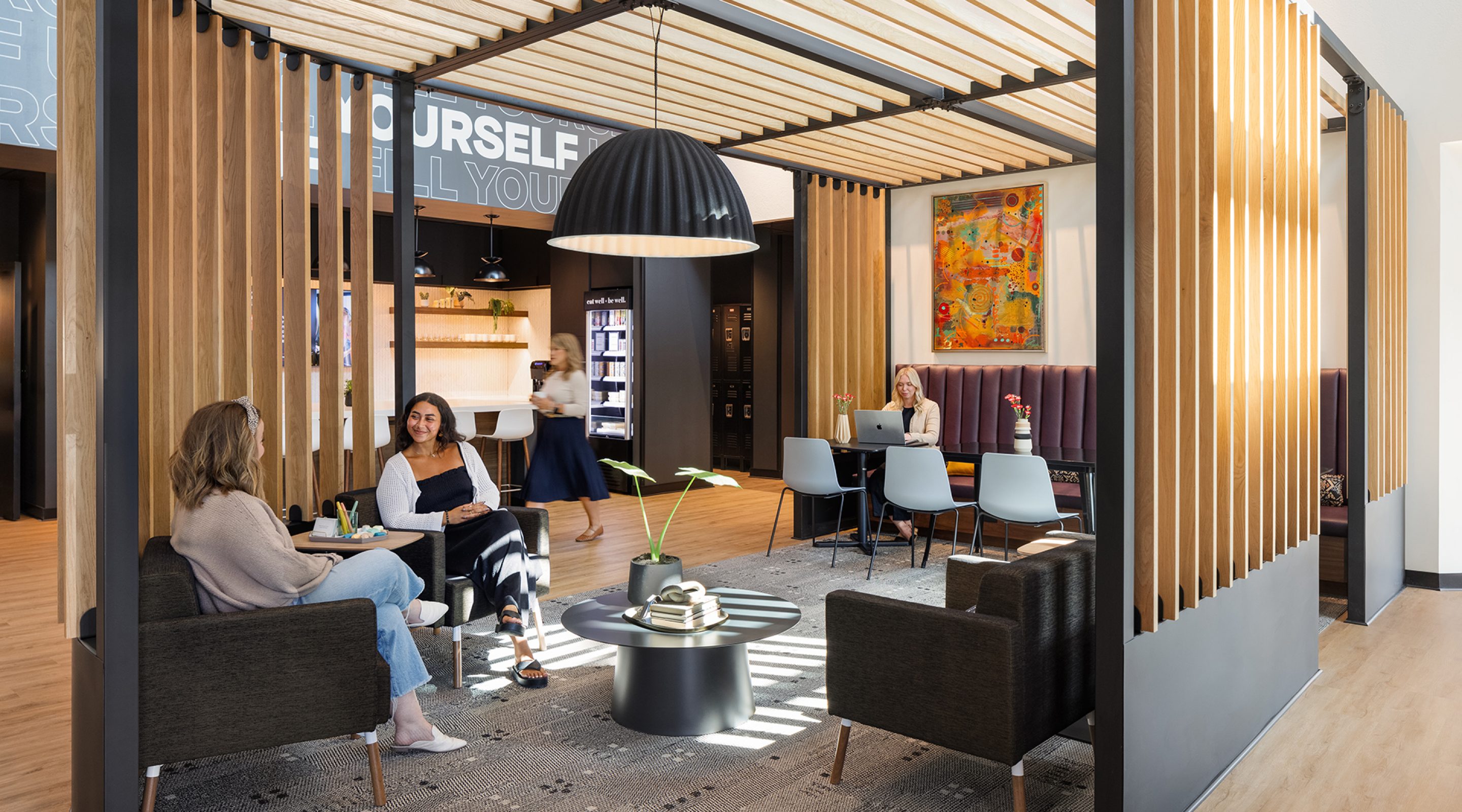 The Lounge at Penzone Base One has a wood slatted structure in the foreground. Inside the structure is a hanging acoustic pendant lamp, dark grey armchairs with two women sitting and a round black coffee table with books and pants on top. At the far end of the structure is a purple banquette with three black tables and grey chairs with a woman sitting and working on her laptop. In the background is the kitchenette with cabinets, chairs and a blonde woman in a blue skirt walking through.