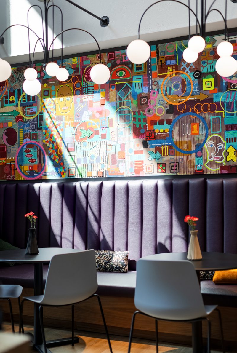 Purple banquette seating at Penzone Base One with light streaming through. There are black round tables in front of the banquette with vases on top and grey chairs. Behind the banquette on the wall is a colorful painting and metal chandeliers above with round glass globes.