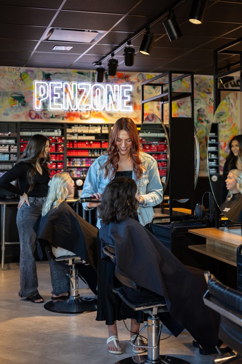 Penzone Base One salon area with two hairstylists and their clients seated in front of mirrors with salon capes on. The stylist's work spaces have a wood table and shelves up above the morrors and are easily movable. In the background is a colorful graffiti-type wall with a large LED sign reading 