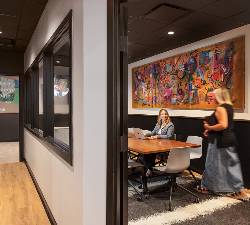 Penzone Base One conference room with two women at a wood table with white rolling chairs. The walls are white with black trim and a colorful painting hangs above their head. The conference room has windows into it from the hallway that leads to the spa treatment area.