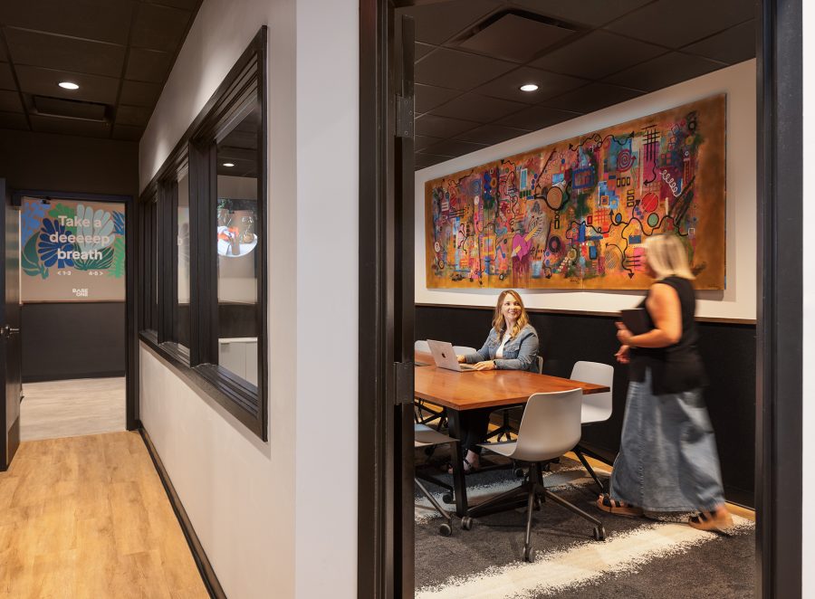 Penzone Base One conference room with two women at a wood table with white rolling chairs. The walls are white with black trim and a colorful painting hangs above their head. The conference room has windows into it from the hallway that leads to the spa treatment area.